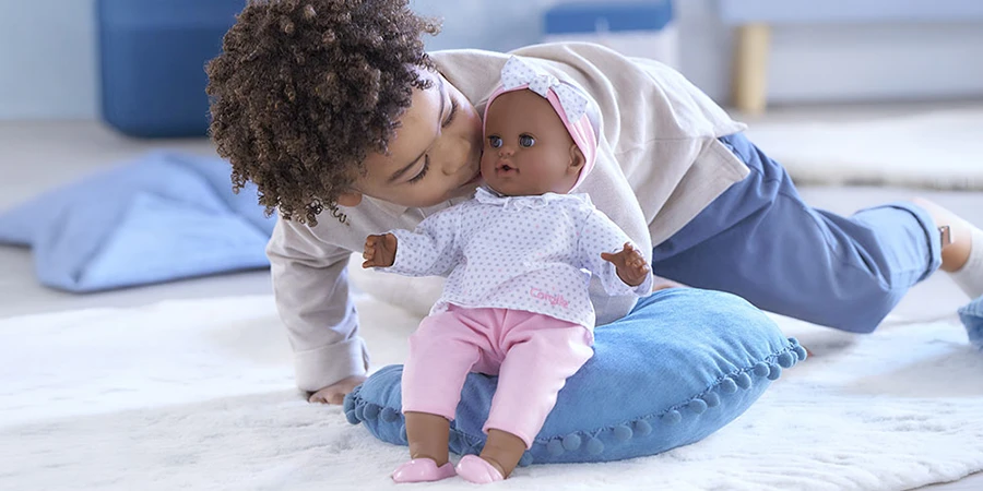 Un enfant embrasse un poupon Corolle habillé en rose et blanc, posé sur un coussin bleu