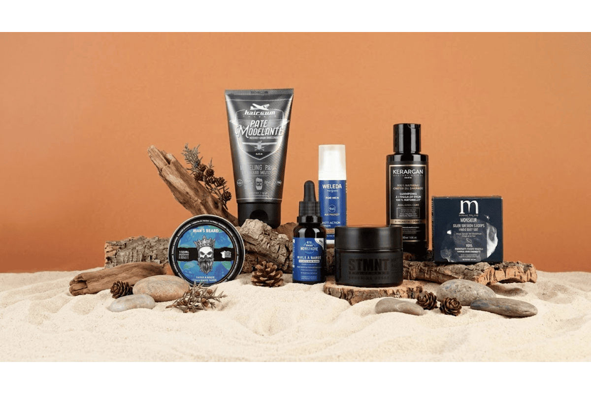 Box Avantages Monsieur avec ses 8 soins disposés sur sable et bois flotté, ambiance plage et nature, avec Big Moustache huile à barbe, Man's Beard baume, STMNT pâte matifiante et Hairgum pâte modelante