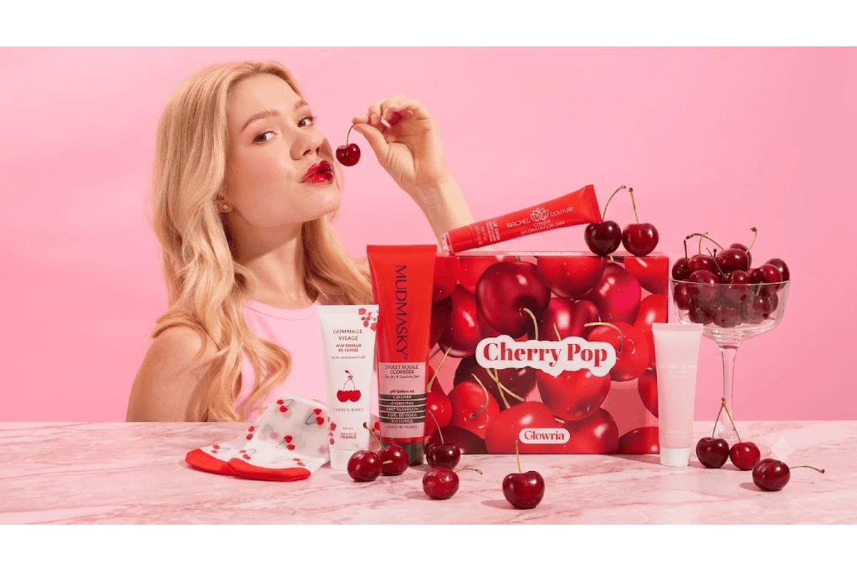 Mannequin blonde avec cerise à la bouche devant la box Glowria Cherry Pop et ses produits Mudmasky, Rachel Couture et Glossy Glam sur fond rose