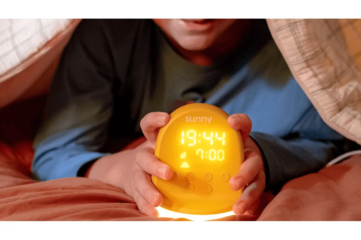 Morphée Sunny réveil enfant orange lumineux affichage 19h44 tenu par un enfant dans son lit sous la couverture ambiance cocon nocturne veilleuse sans écran test produit gratuit Mamadvisor