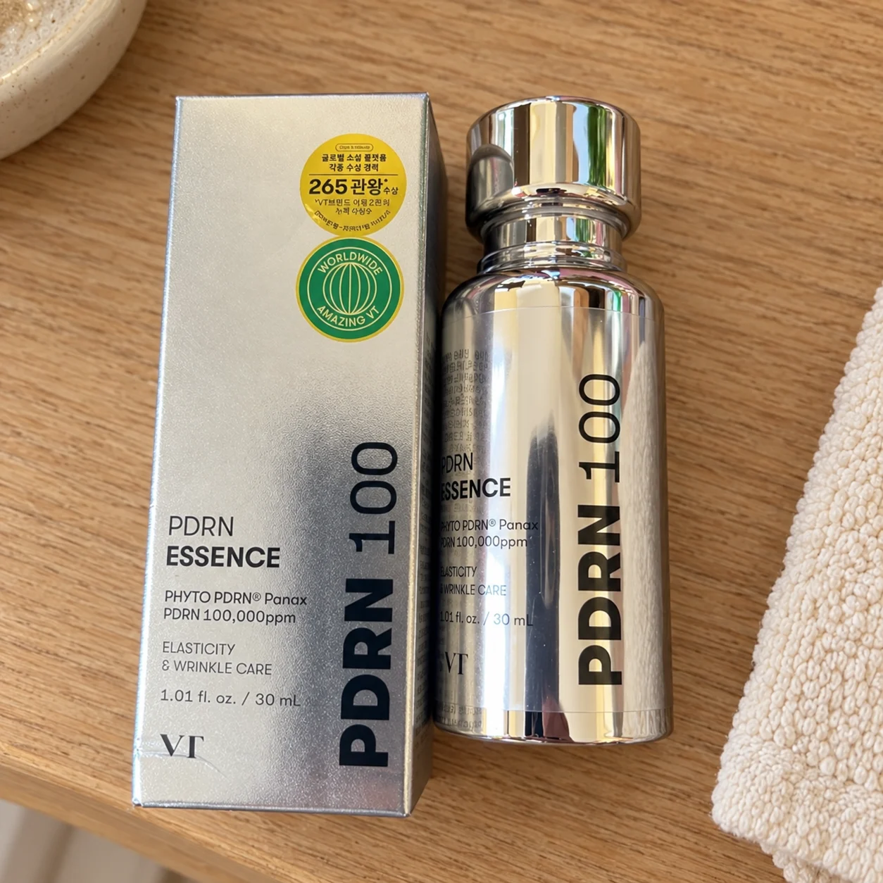Flacon et boîte argentés de la VT PDRN Essence 100 posés sur une tablette en chêne clair
